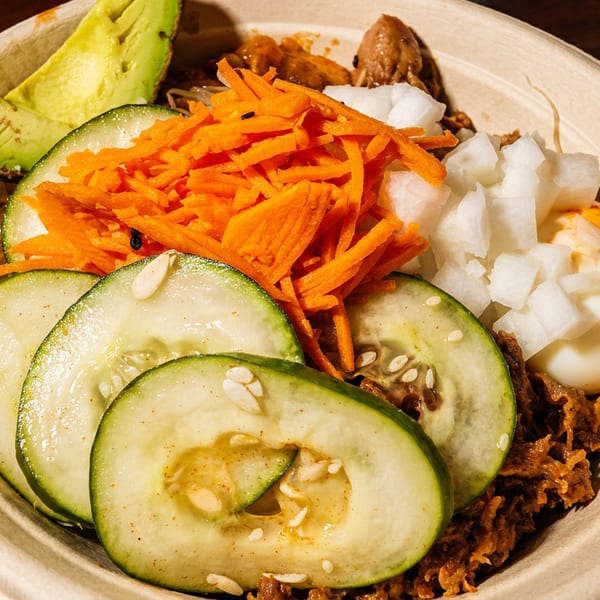 Close-up of a bowl with sliced cucumbers, shredded carrots, avocado, and diced onions.